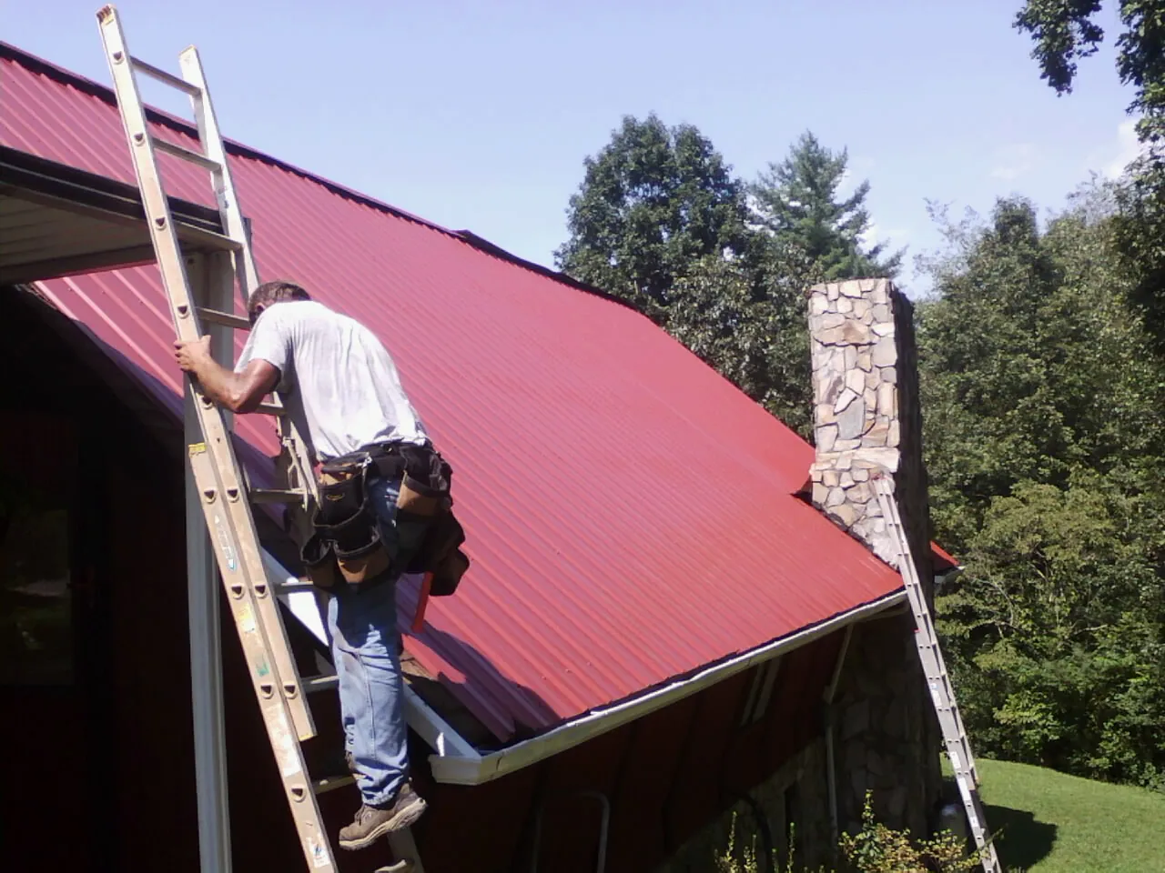 Roofer on a ladder beside a red metal roof and stone chimney — Soffit & Fascia Repair by Roofing Brothers in Rock Springs